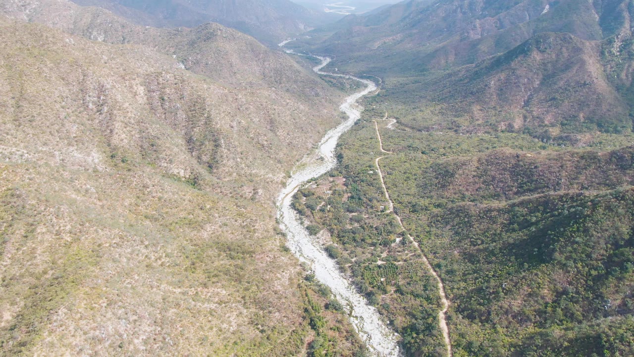 Aerial frame in San Dionisio, Baja California Sur, with the river bending through the center-left valley floor, a thin dirt road nearby, dry hills on both sides, and no visible people.