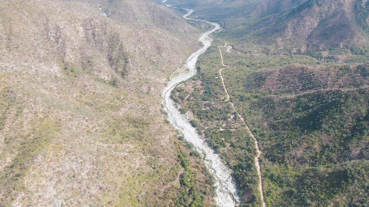 Aerial frame in San Dionisio, Baja California Sur showing mixed dry and green terrain around a winding river, with the dirt road visible along the valley and no visible people.