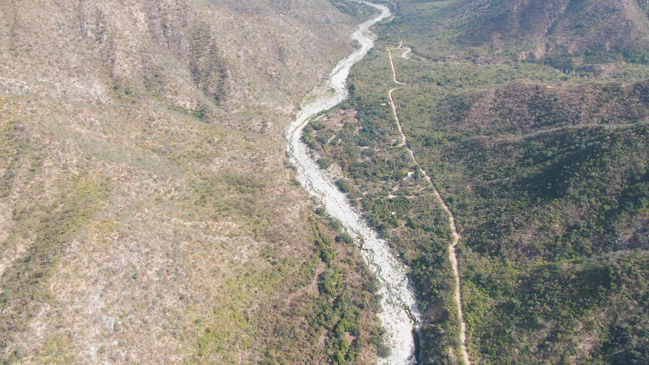 Aerial frame from San Dionisio, Baja California Sur, showing a pale river curving through a mountain valley with dry tan slope at left, darker forest at right, and no visible people.