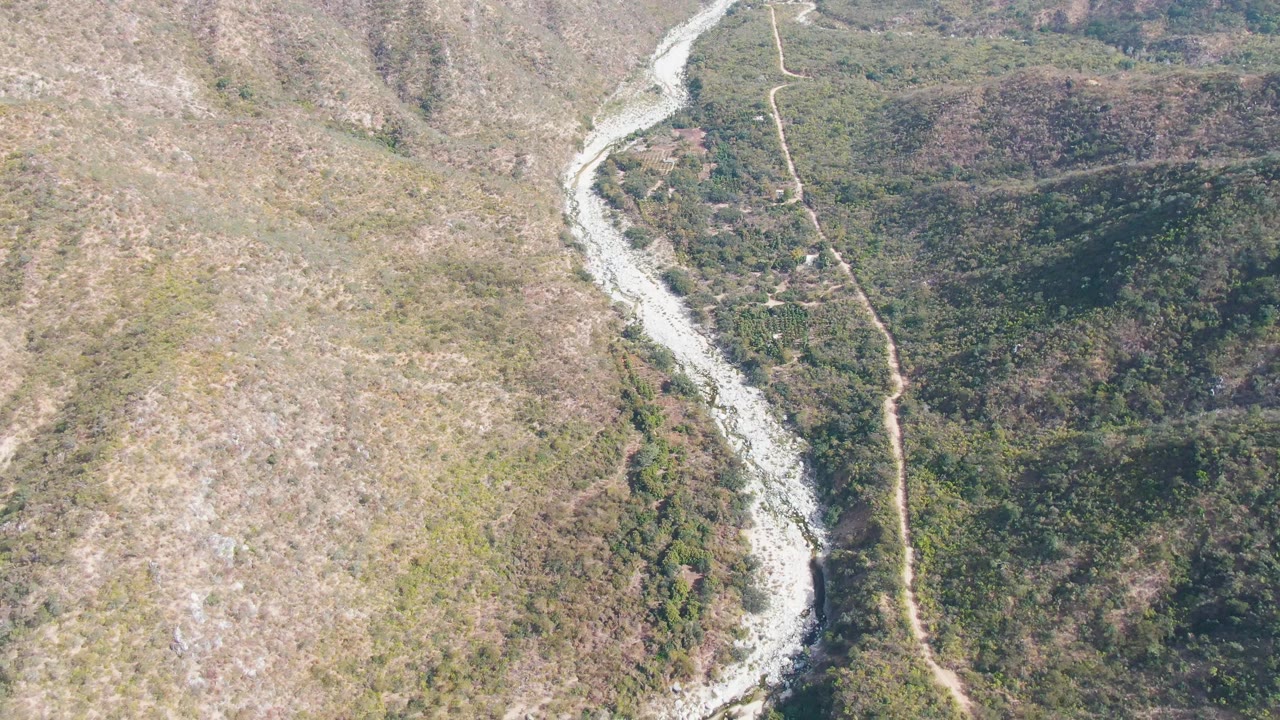 San Dionisio aerial frame showing a bright pale river bend bordered by dense green vegetation on the right side of the valley, drier rocky slope on the left, and no visible people.