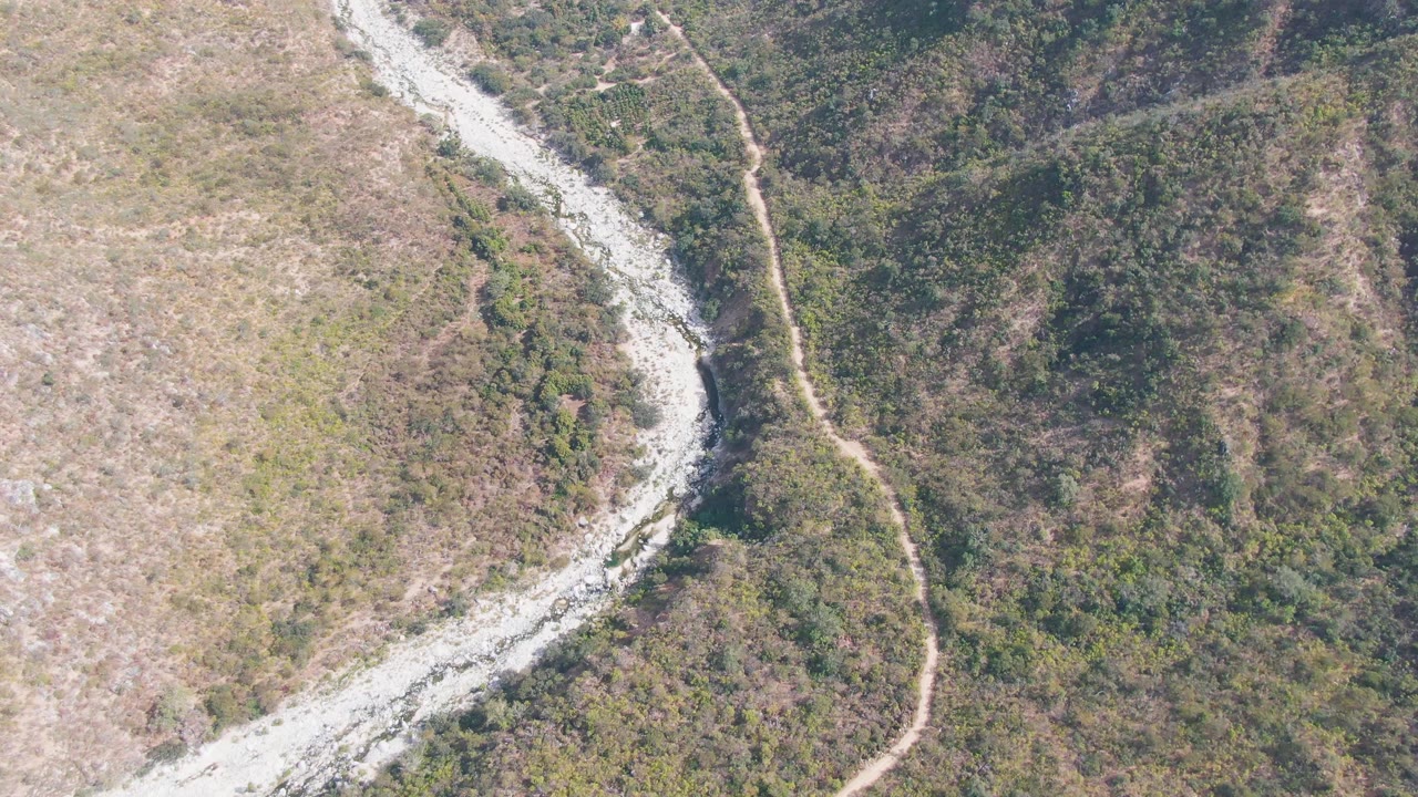 At 0:01, the San Dionisio aerial view keeps the curving river channel near the left side while the dirt track runs through green and tan hillside vegetation with no visible people.