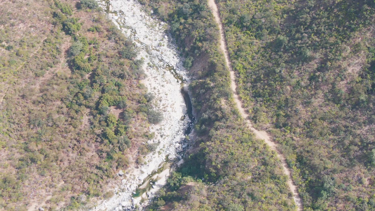 Aerial view in San Dionisio, Baja California Sur, showing a pale rocky riverbed curving past green and brown hillside vegetation with a dirt path on the right and no visible people.