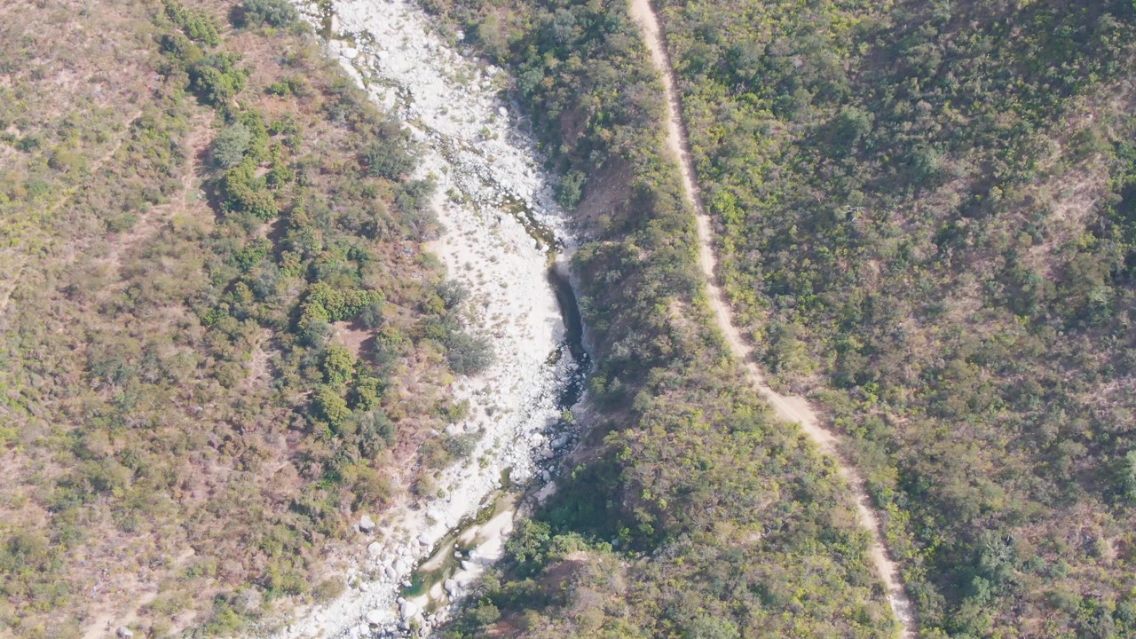 Aerial frame from San Dionisio, Baja California Sur, with the rocky river channel near the left side, a narrow dirt path on the right, mixed green brush, dry brown ground, and no visible people.
