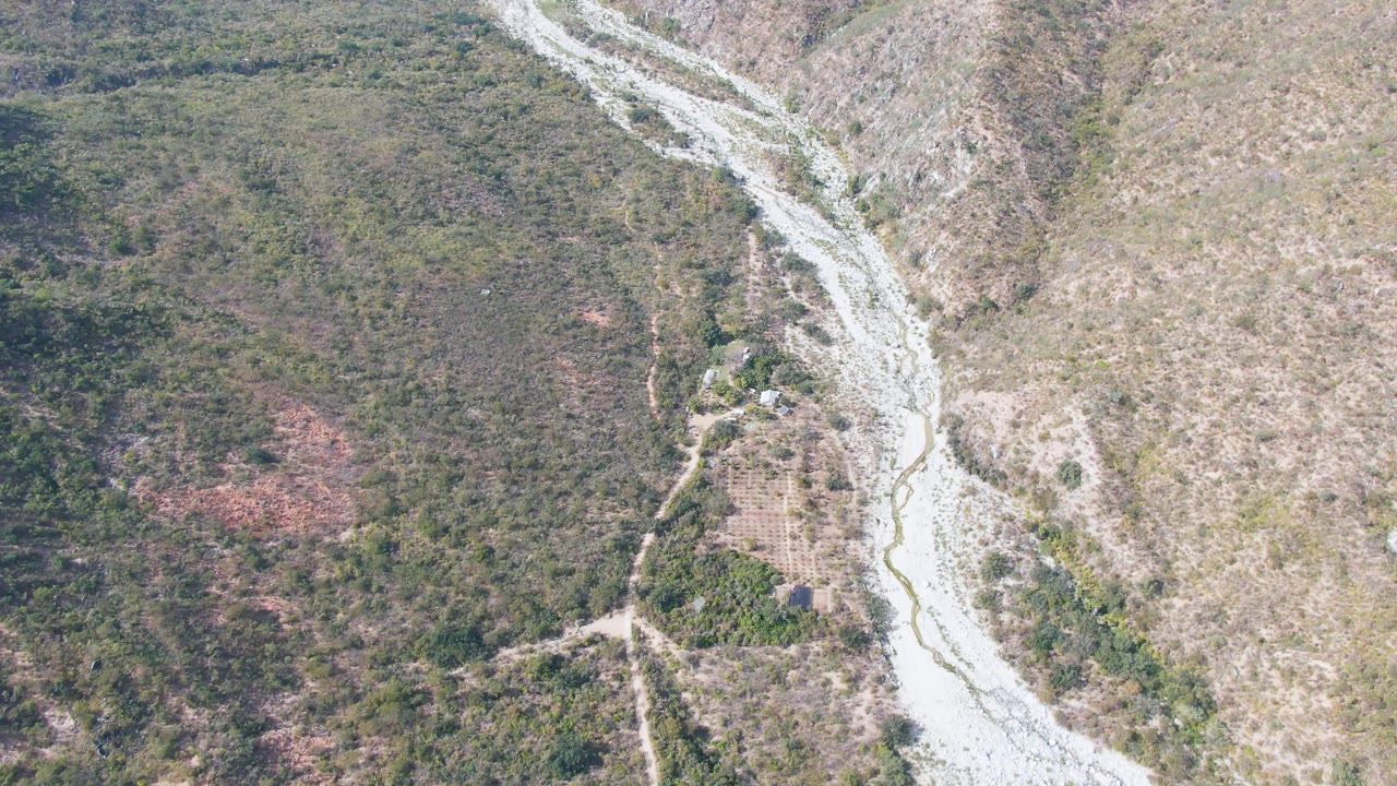 Aerial frame over San Dionisio, Baja California Sur, showing a pale riverbed cutting diagonally between green vegetation and brown dry slopes, with no visible people.