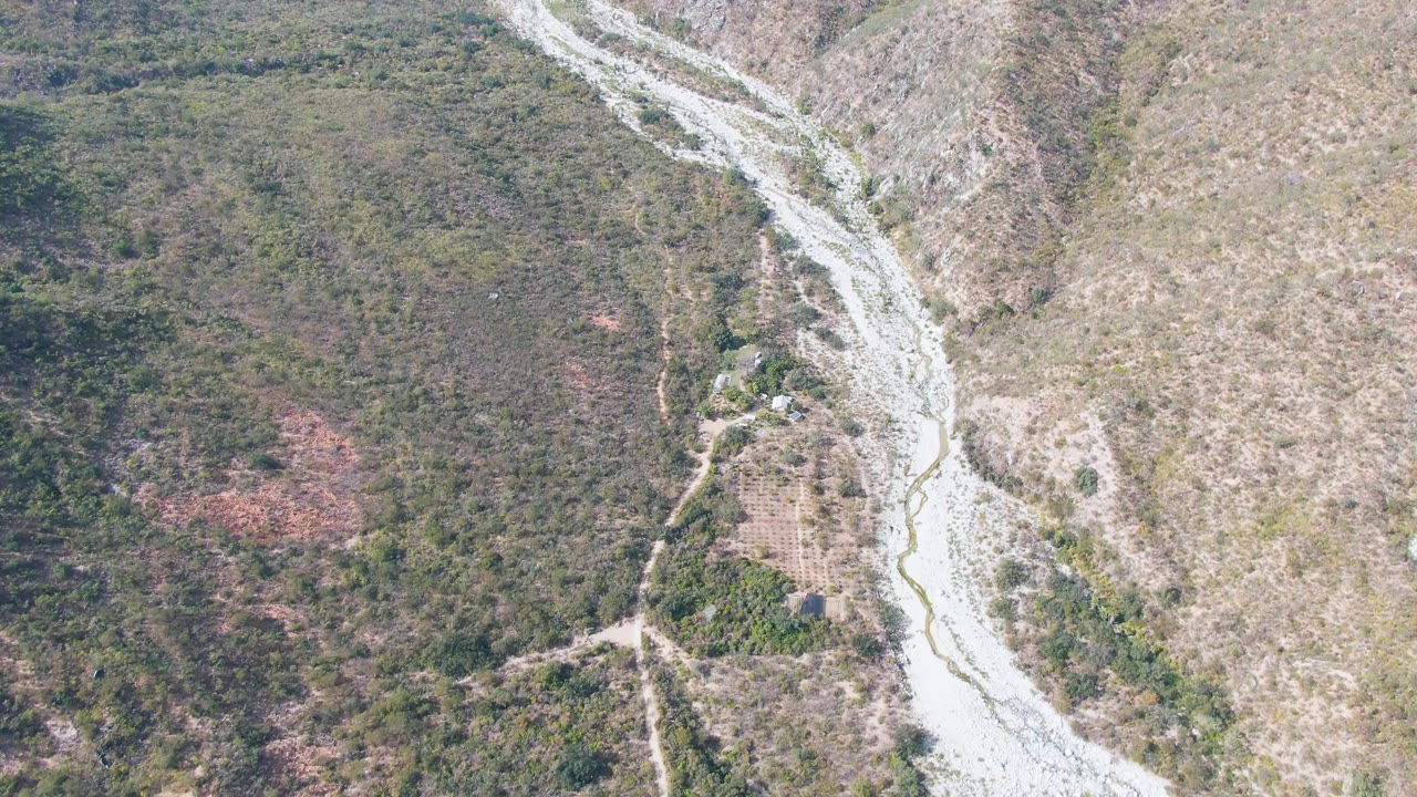 High aerial frame in San Dionisio, Baja California Sur, with the riverbed near the right side, scrubby green terrain to the left, dry hillside to the right, and no visible people.