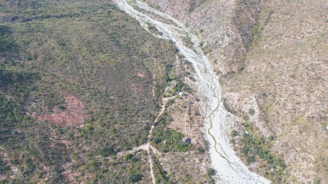 Drone-style overhead frame from San Dionisio, Baja California Sur, showing the bright riverbed bending through rugged hills with patches of green brush and no visible people.