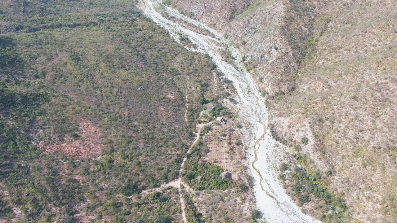 Aerial frame over San Dionisio, Baja California Sur, showing the riverbed shifted across the frame with brushy hillside texture, dry open slope, faint dirt tracks, and no visible people.