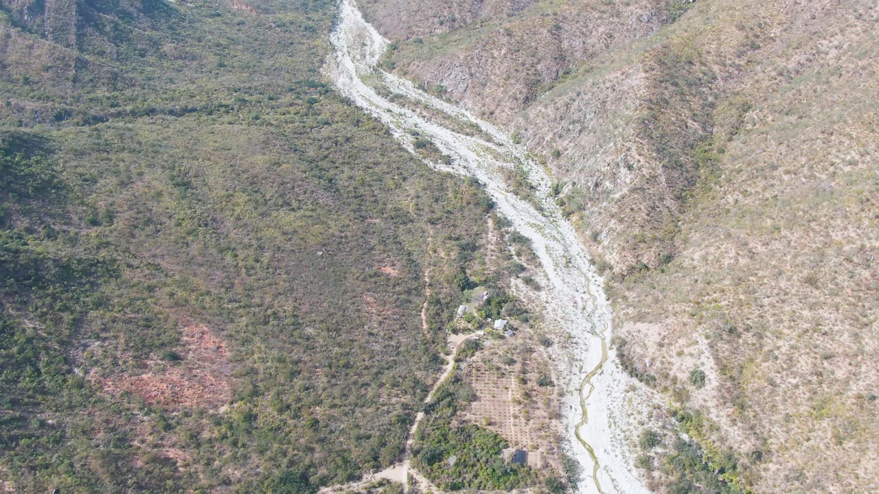 High aerial frame from San Dionisio, Baja California Sur, showing the riverbed centered between green-brown slopes, dirt paths, and no visible people.