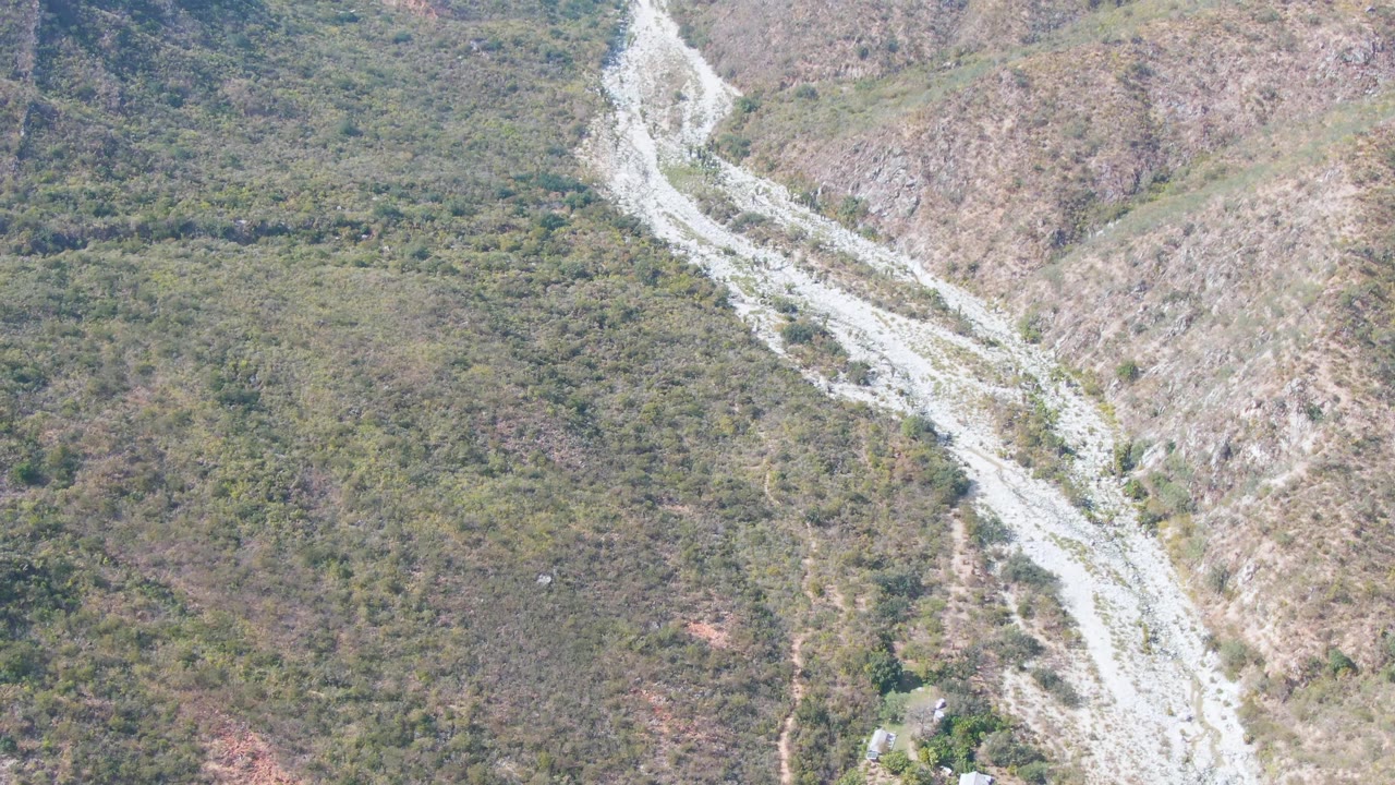 Overhead view in San Dionisio, Baja California Sur with a bright rocky road or wash line bending through low vegetation and exposed hillside, with no visible people.