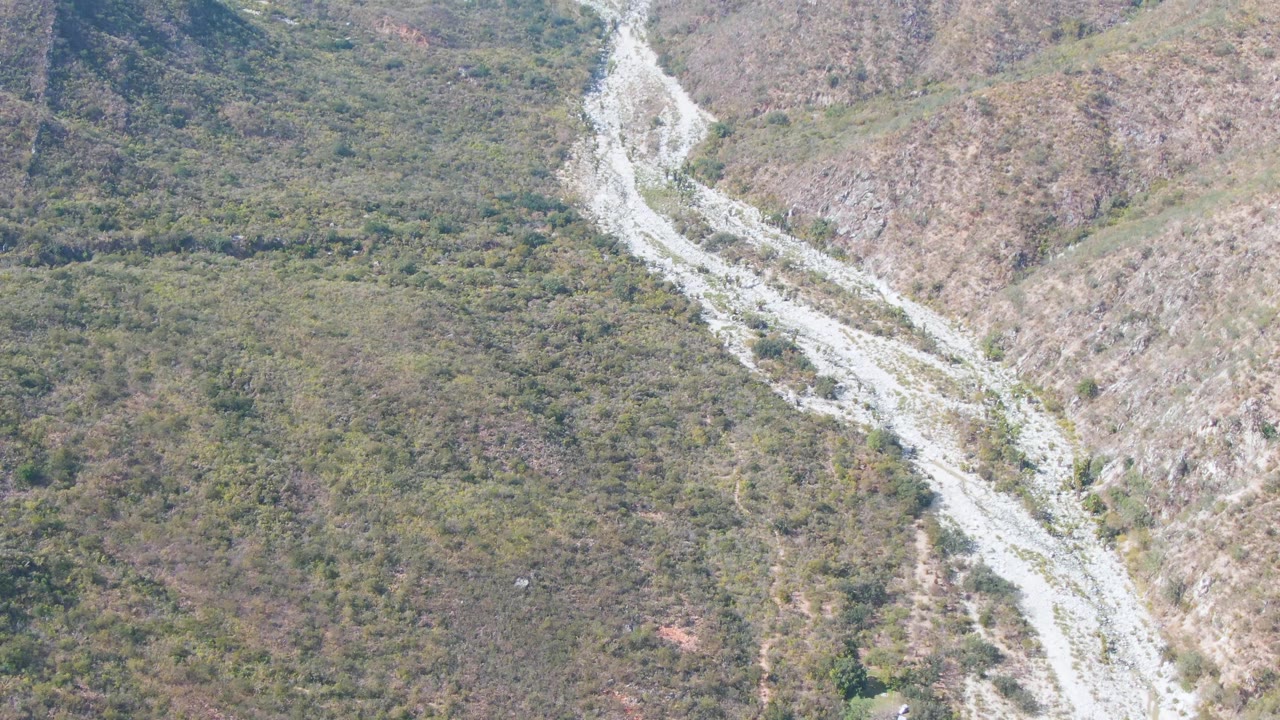 Drone-like view of a narrow pale track cutting through a vegetated mountain valley.