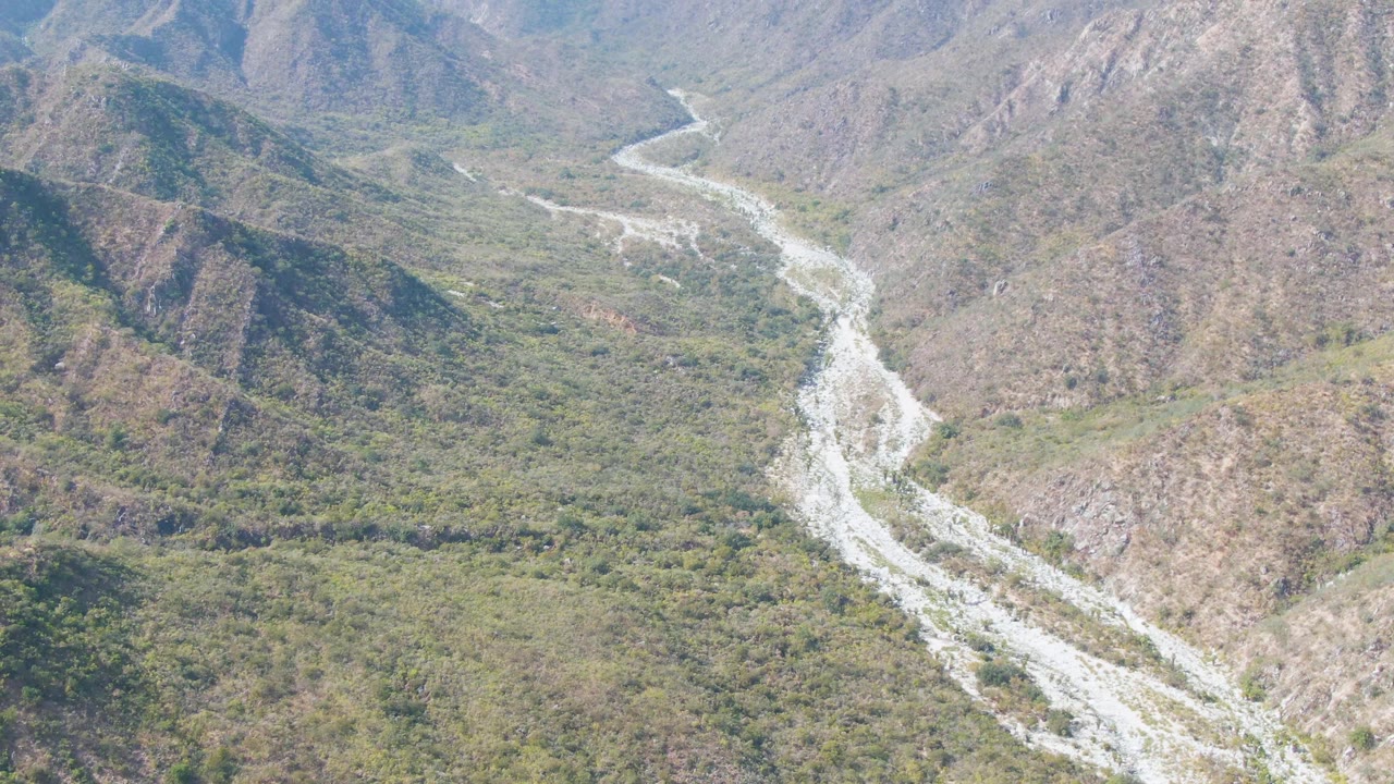 Aerial frame from San Dionisio, Baja California Sur, with the river channel curving through a green valley floor and rocky brown hillsides, no visible people.