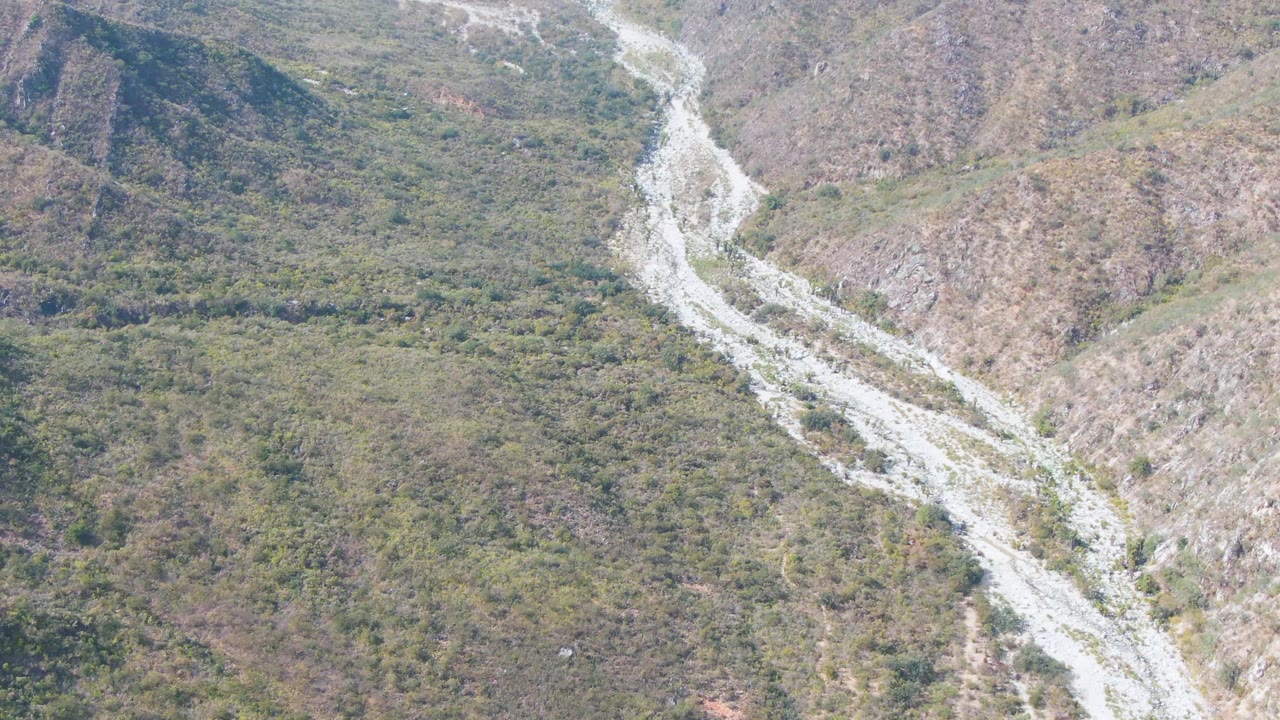 Aerial view of a pale winding riverbed between scrub-covered mountain slopes.