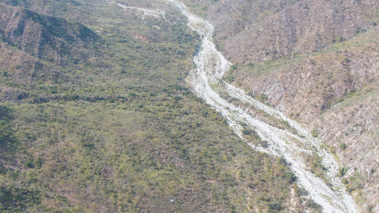 Aerial frame from San Dionisio, Baja California Sur, with the rocky drainage channel more centered between uneven vegetated ridges and no visible people.