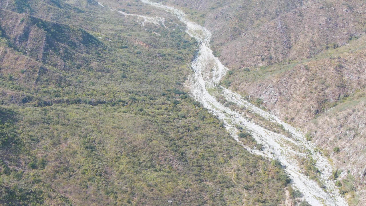 Aerial frame from San Dionisio, Baja California Sur, showing the pale winding riverbed beside patchy green valley vegetation and brown hillsides with no visible people.