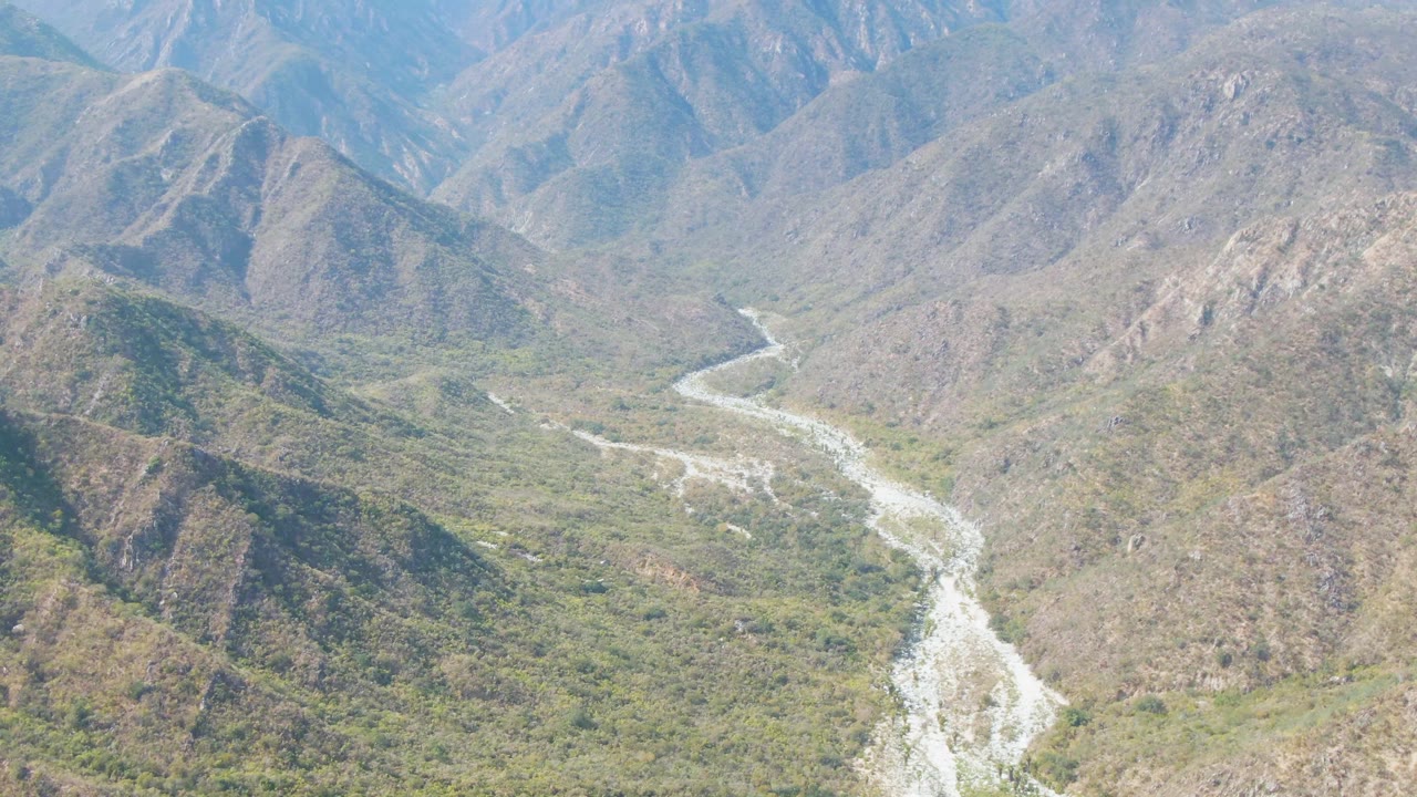 San Dionisio aerial view with the river channel slightly more centered between scrub-covered slopes and hazy mountain ridges, with no visible people.