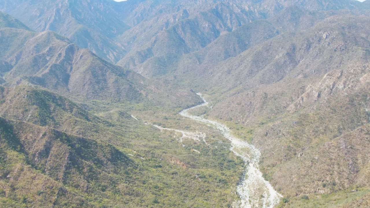 Aerial view in San Dionisio showing a bright riverbed snaking away from the foreground valley floor toward layered blue-gray ridges, with no visible people.