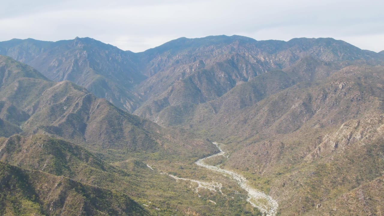 Aerial view over San Dionisio, Baja California Sur, showing a pale winding riverbed cutting through green and brown rocky hills with layered mountains beyond and no visible people.