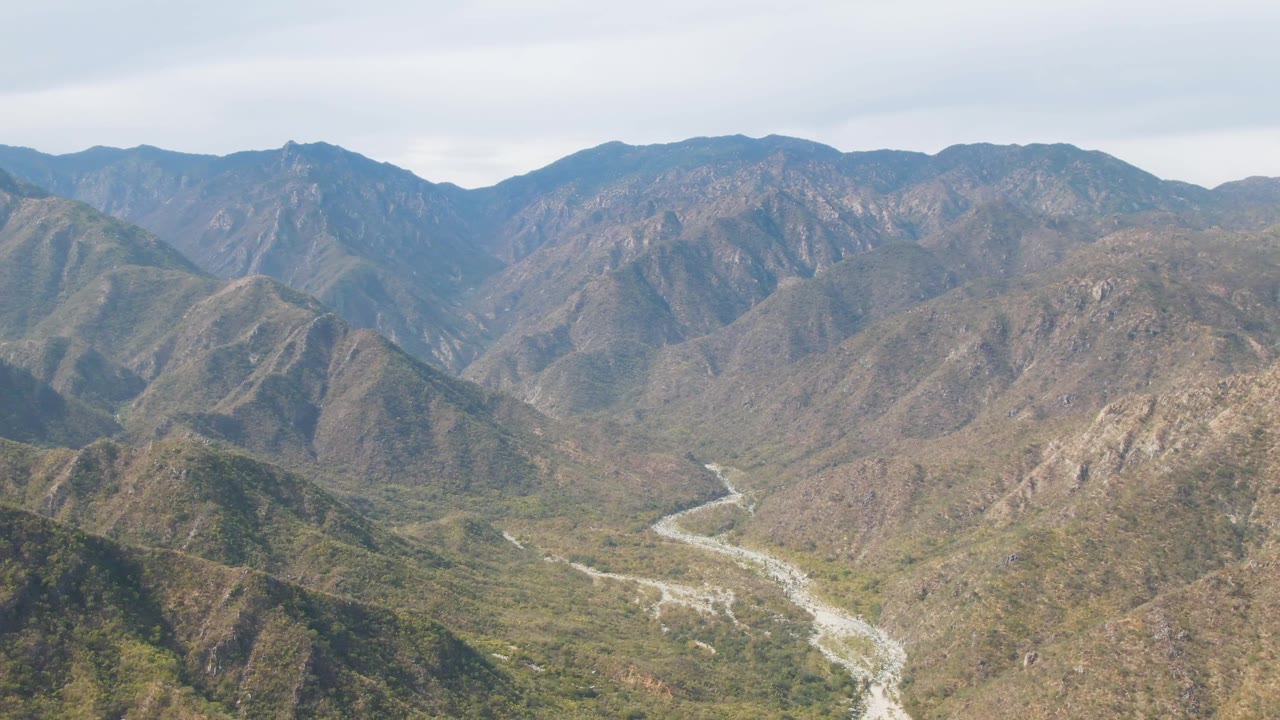 Aerial view in San Dionisio, Baja California Sur, showing rocky foreground hills, a pale riverbed bending through the valley floor, muted green vegetation, and no visible people.