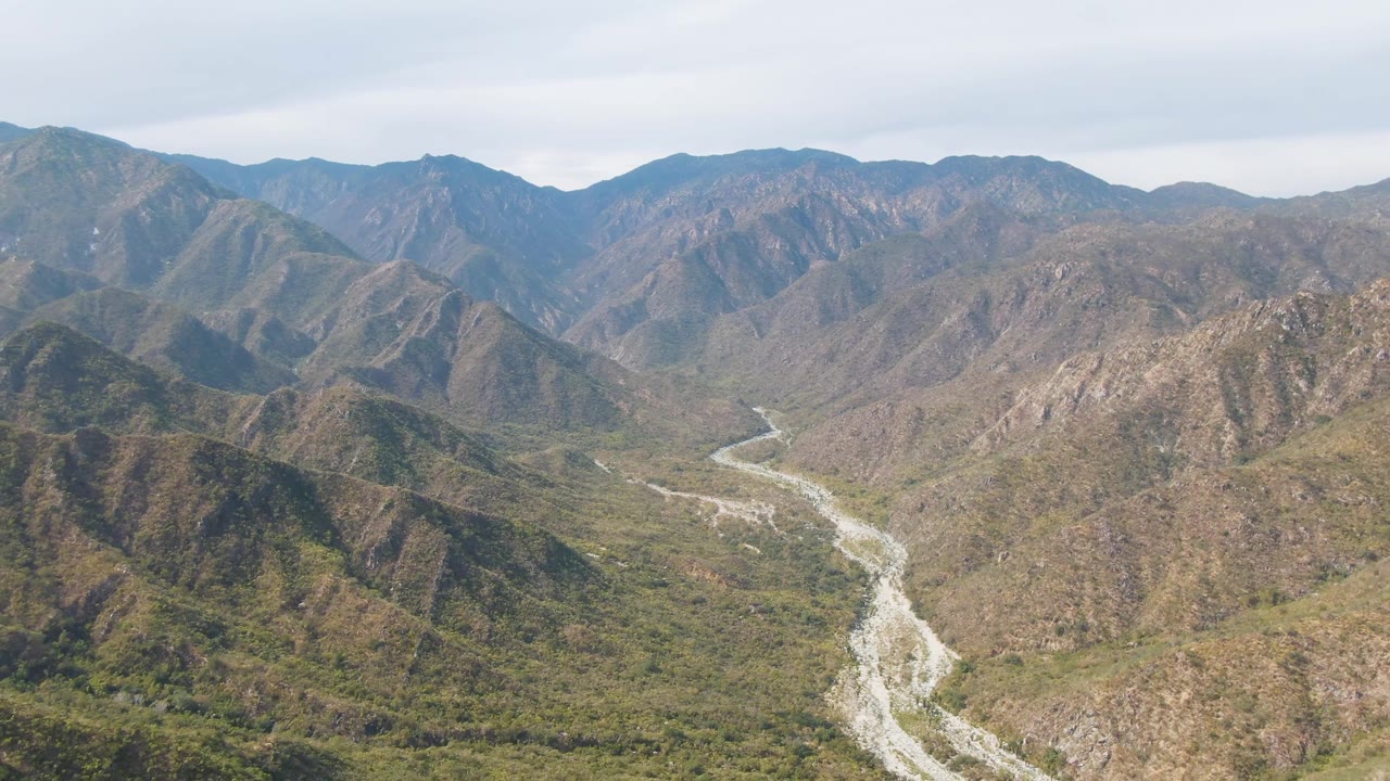 Aerial view over San Dionisio, Baja California Sur, with a pale winding riverbed crossing a green and brown mountain valley beneath a gray sky, no visible people.