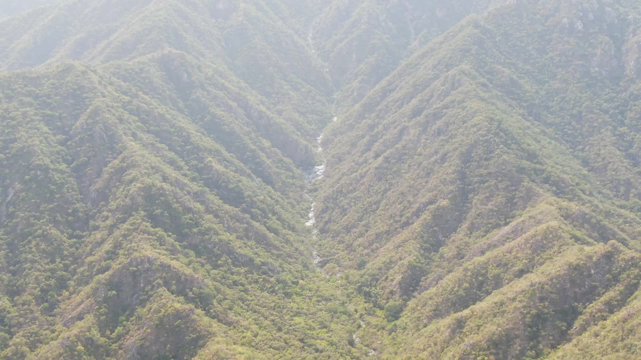 High aerial frame over San Dionisio ridges, with dense green vegetation across the slopes, pale haze in the distance, and no visible people.