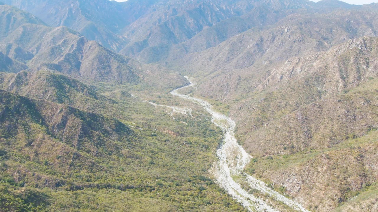 Aerial frame of San Dionisio, Baja California Sur, showing the riverbed curving through a broad valley, dry ridges on both sides, and no visible people.
