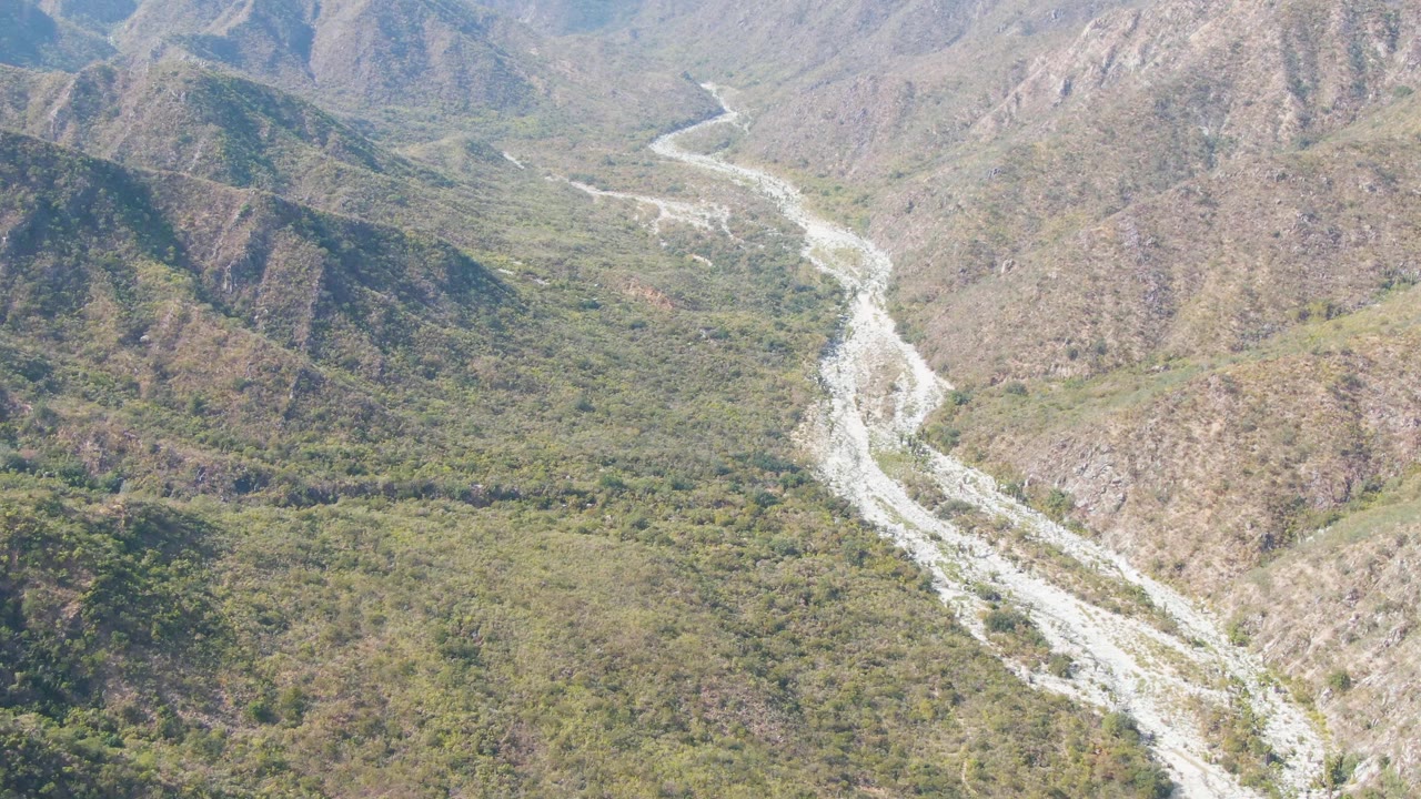 Aerial frame showing the river bend farther along the valley floor in San Dionisio, with scrub vegetation, exposed rock, hazy daylight, and no visible people.