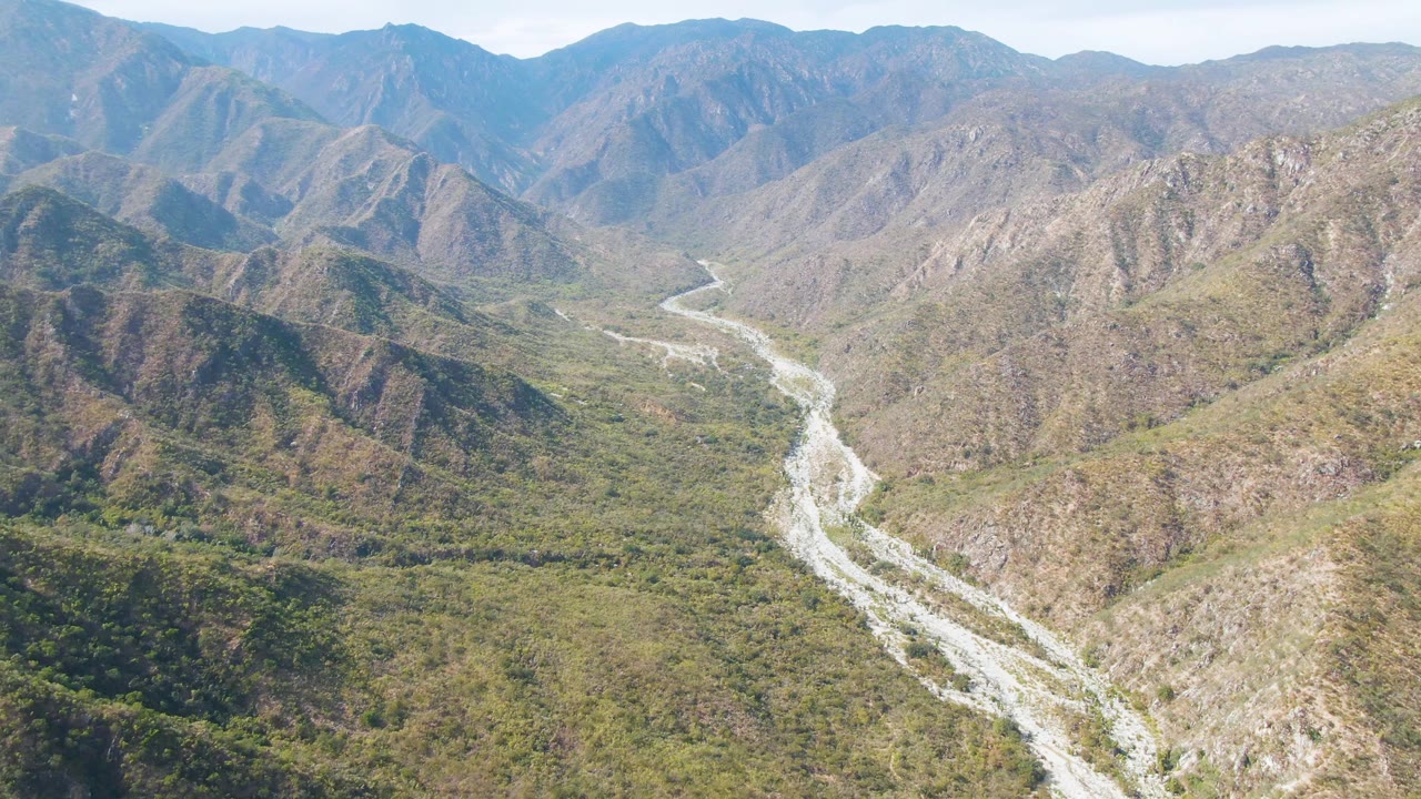 Opening aerial frame over San Dionisio, Baja California Sur, with a pale winding riverbed below rocky green-brown ridges and no visible people.