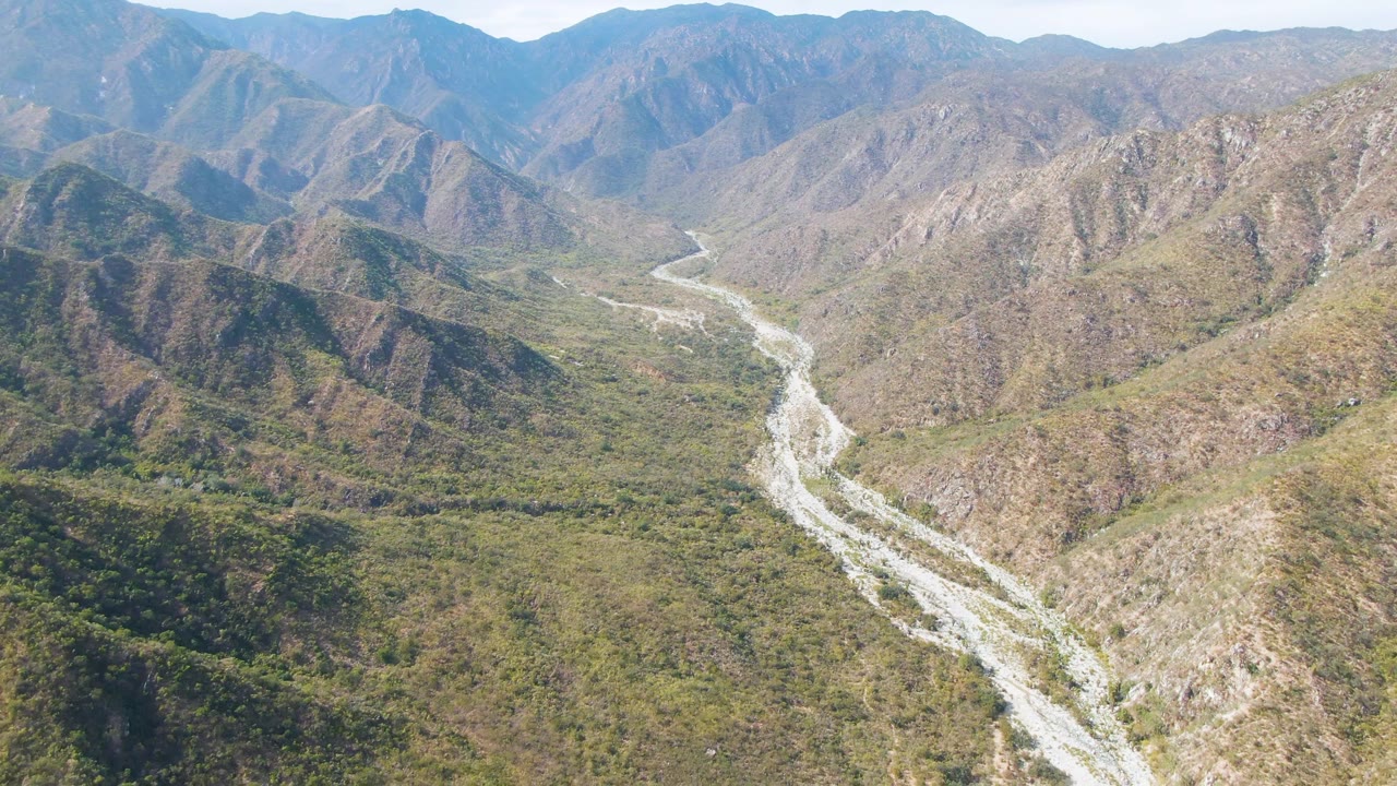 Mid-clip aerial frame over San Dionisio, Baja California Sur, with the pale riverbed threading between rugged hills and a hazy line of distant mountains, no visible people.