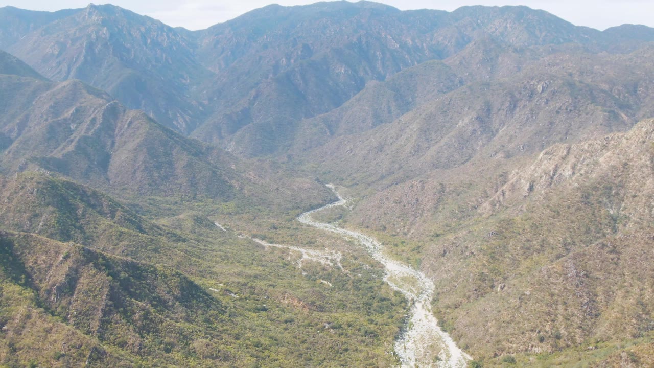 Mid-clip aerial view of San Dionisio terrain, with the pale river bending through the valley and layered rocky mountains filling the background with no visible people.