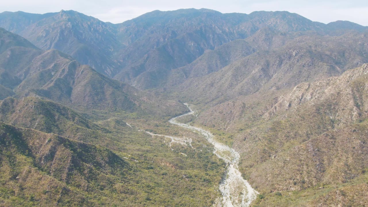 Aerial frame in San Dionisio showing rocky foreground hills, a winding river below, and blue-gray mountain ridges across the horizon with no visible people.