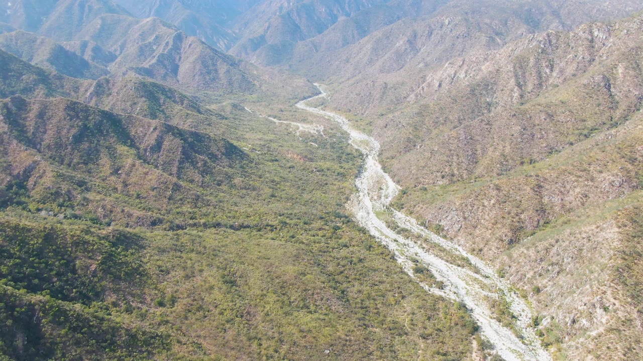 Aerial frame from San Dionisio, Baja California Sur showing a pale winding river between green valley vegetation and rocky mountain slopes, with no visible people.