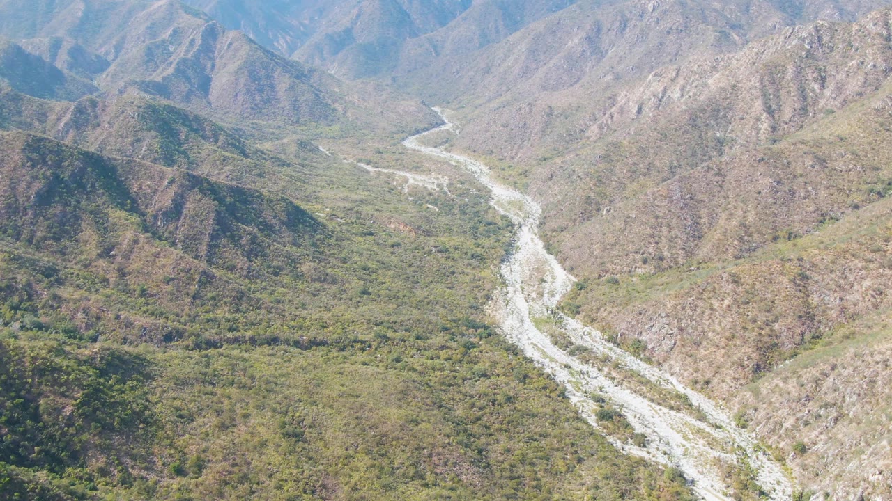 Drone-height view over San Dionisio, Baja California Sur with a pale river channel bending through mixed green vegetation and exposed rocky slopes, no visible people.