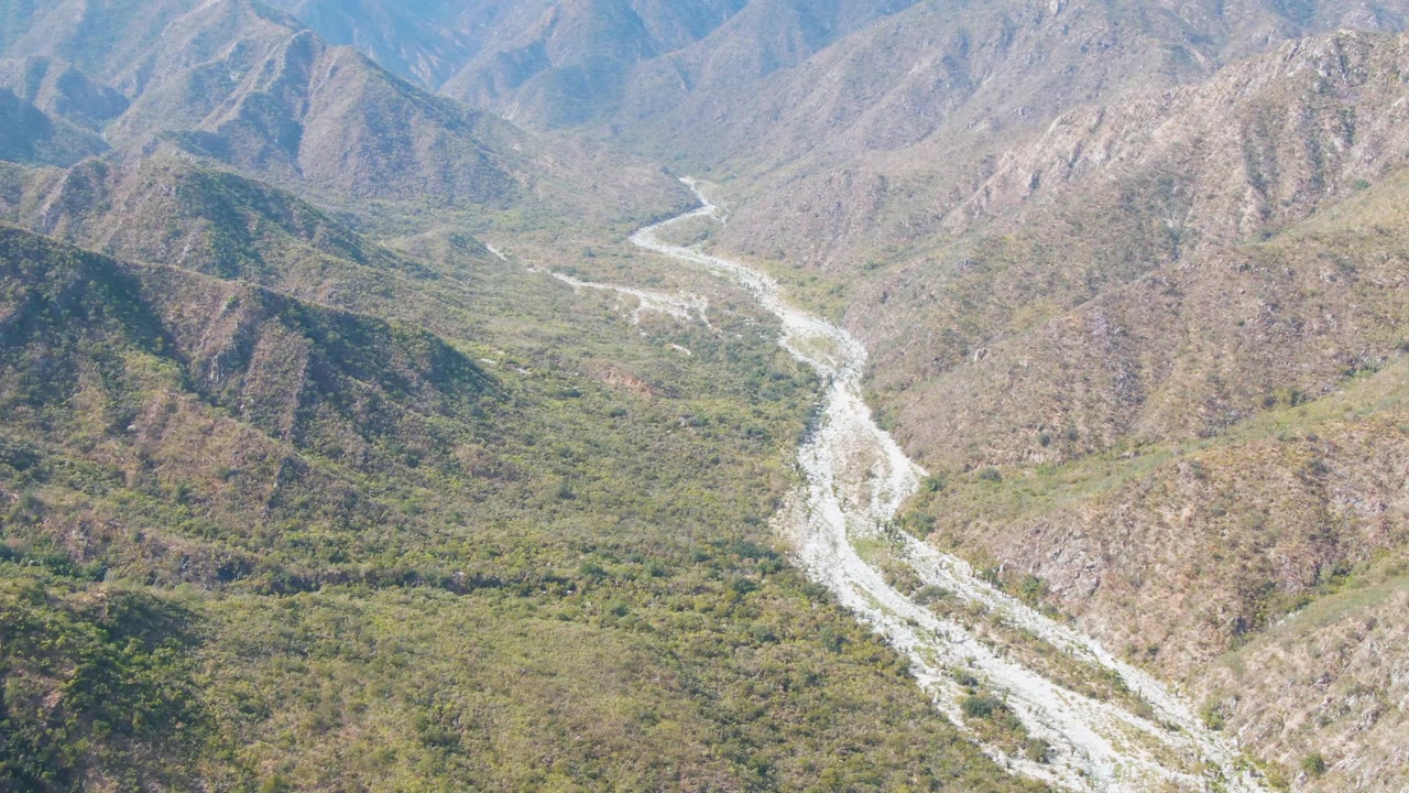 Aerial landscape from San Dionisio, Baja California Sur showing the river winding between rugged hills, green patches, and hazy mountain background, with no visible people.