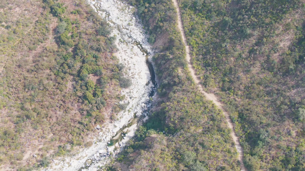 One second into the San Dionisio clip, the rocky river channel bends through mixed dry brush and greener shrubs while the dirt track stays near the right side, with no visible people.