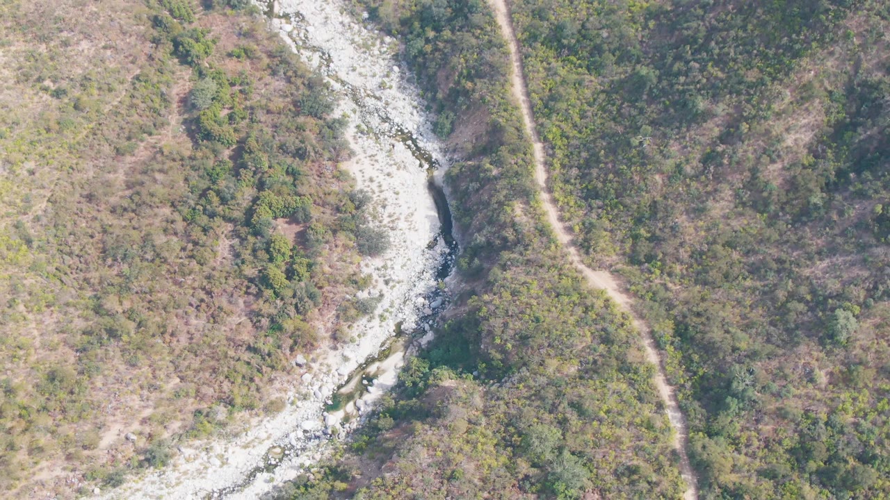 At 0:02, the San Dionisio aerial view holds on the winding rocky streambed with the narrow dirt path more vertical along the right side and no visible people.