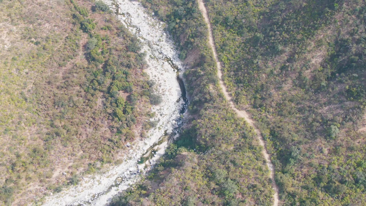 Midway through the clip, the San Dionisio frame shows the pale wash curving through dry hillside brush with denser green vegetation on one side and no visible people.