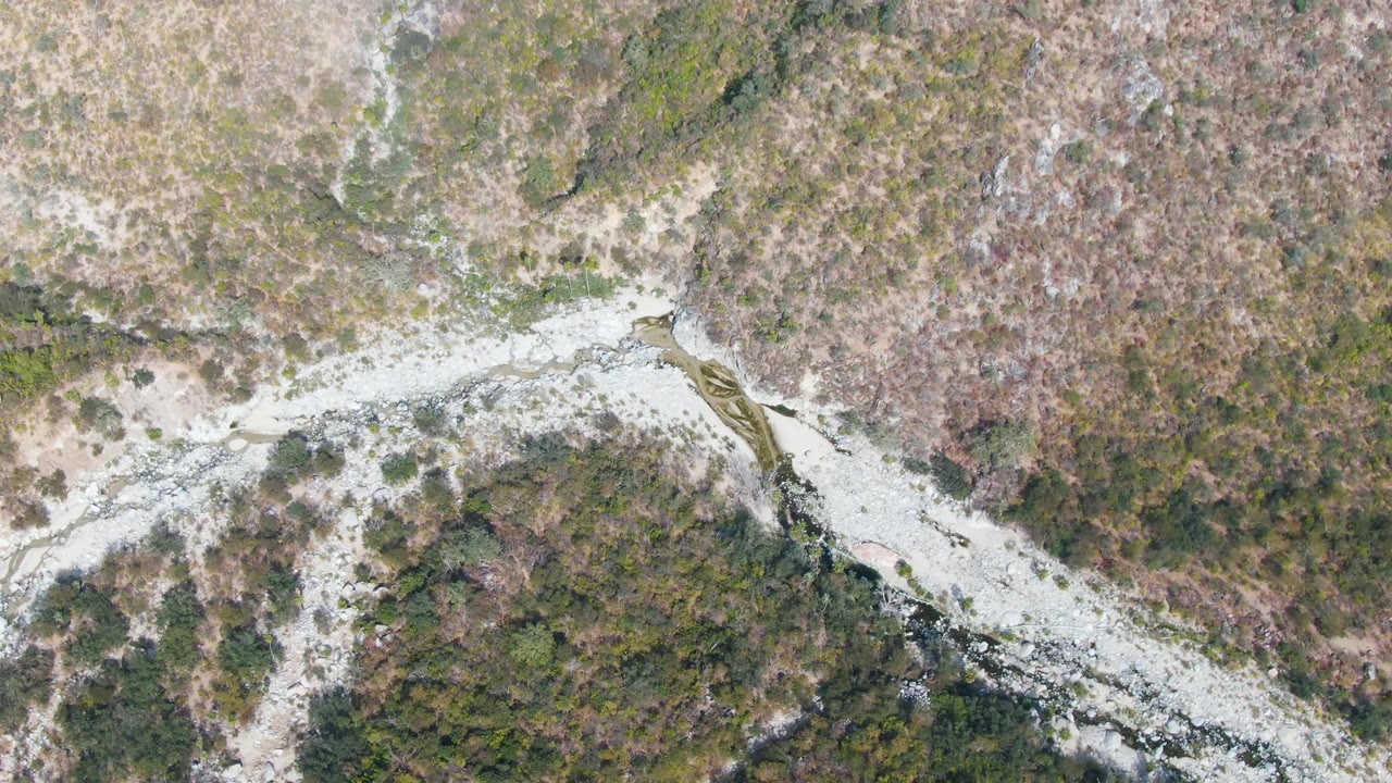Overhead San Dionisio view with a rocky stream bend, small frothy sections in the water, and mixed green and brown terrain around it, no visible people.