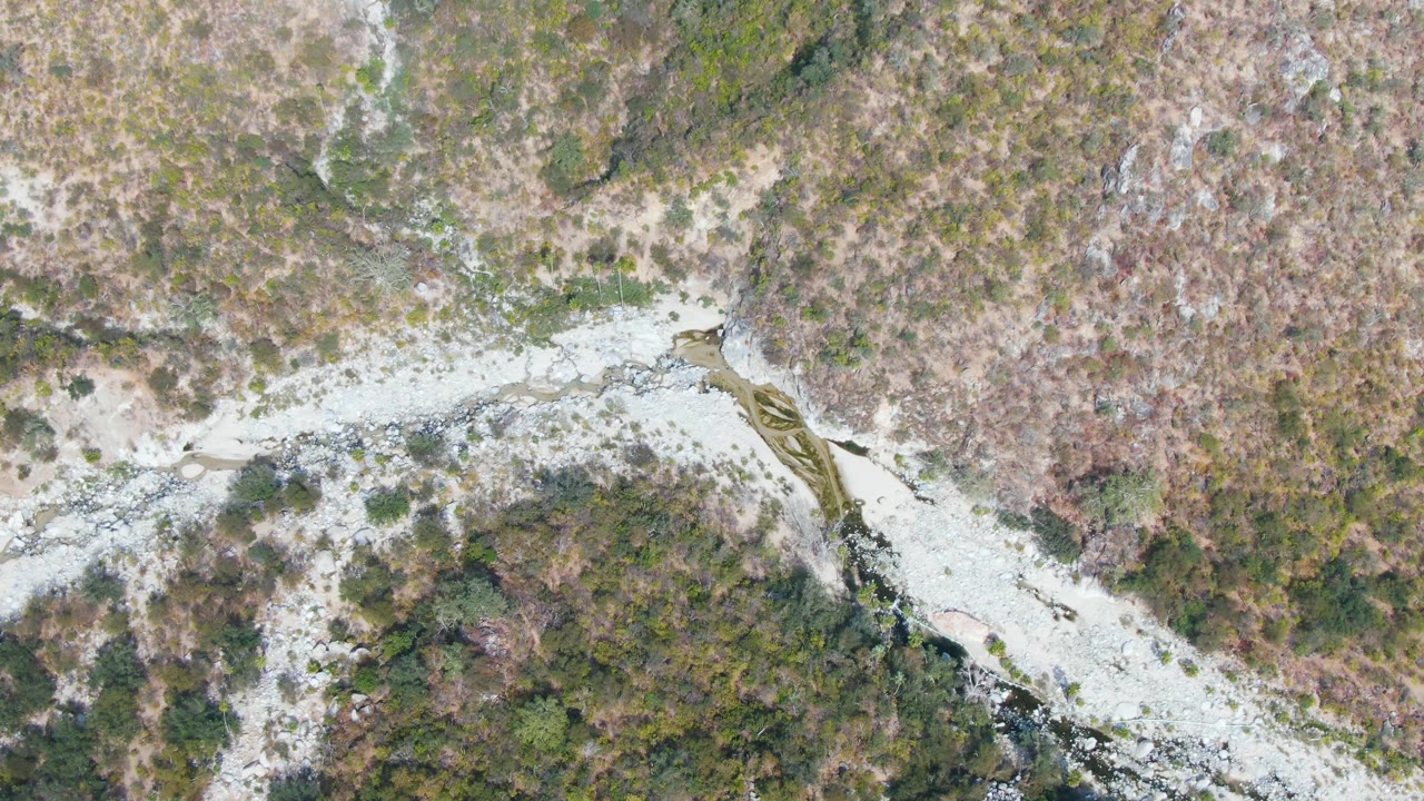 Top-down San Dionisio frame where the pale riverbed runs between rocky banks and brush-covered mountain ground, with no visible people.