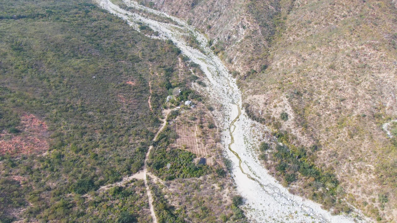 Aerial San Dionisio frame showing a pale river wash at center, branching dirt paths near small cultivated plots, surrounding scrub, and no visible people.