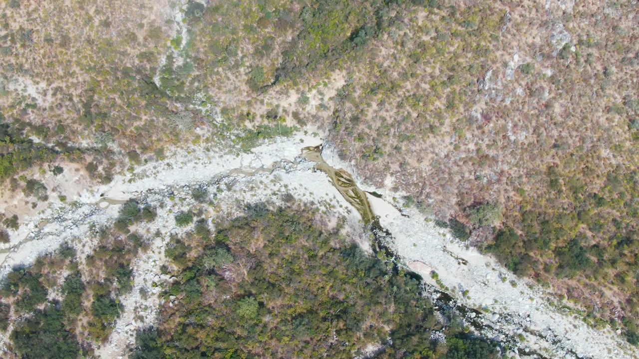 Top-down San Dionisio frame showing the rocky watercourse stretching across the image, bordered by dry brush, green patches, and no visible people.