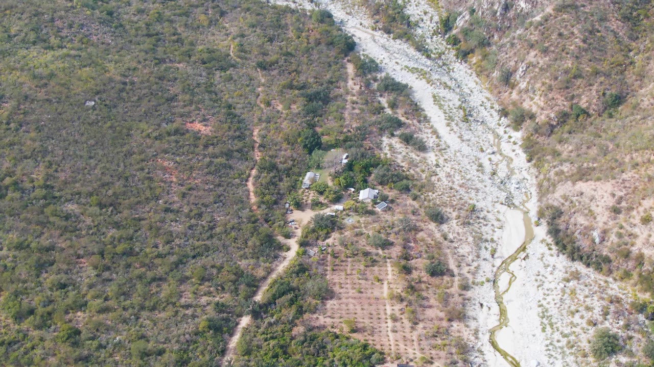Aerial frame over San Dionisio, Baja California Sur showing a pale rocky riverbed, thin muddy current, scrub forest, dry farm rows, dirt tracks, small buildings, and no visible people.