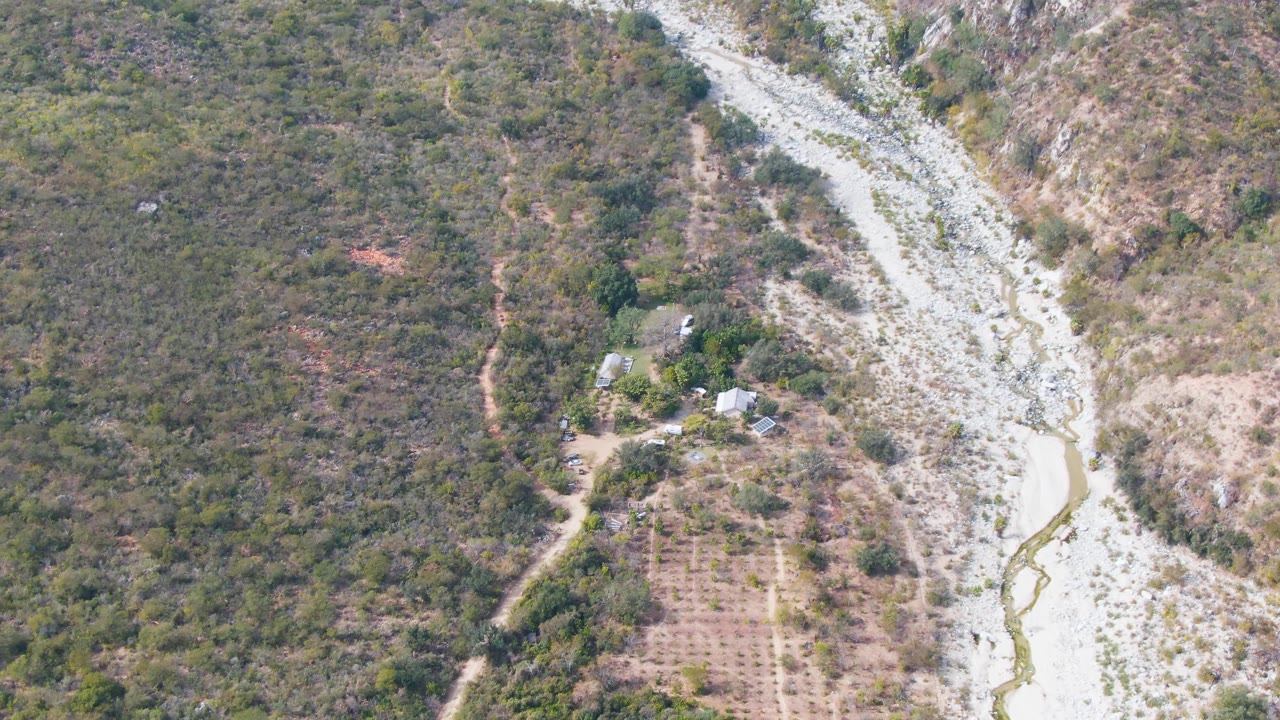 Second aerial frame in San Dionisio, Baja California Sur with the stony river channel running along the right side, patchy green-brown vegetation, farm rows, small structures, and no visible people.