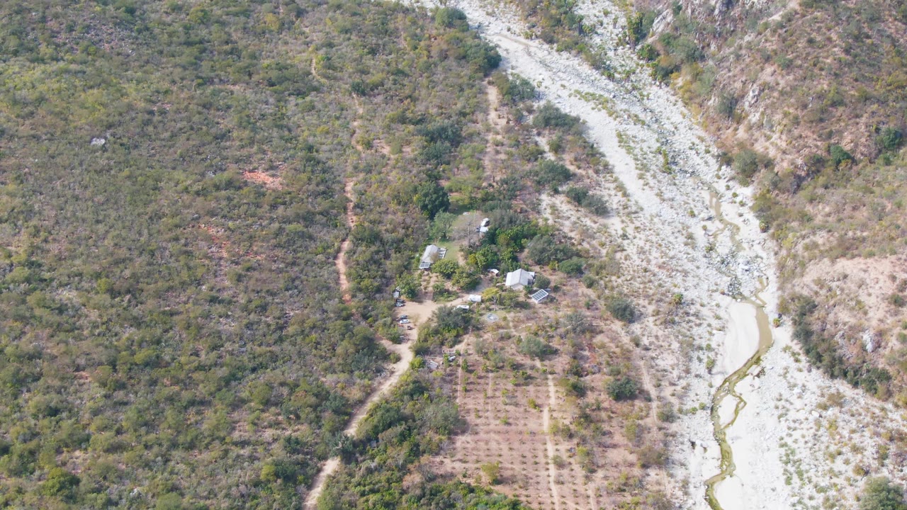 Aerial frame over San Dionisio, Baja California Sur, showing a pale rocky riverbed, scrubby hills, dirt paths, small buildings, a cultivated field, and no visible people.