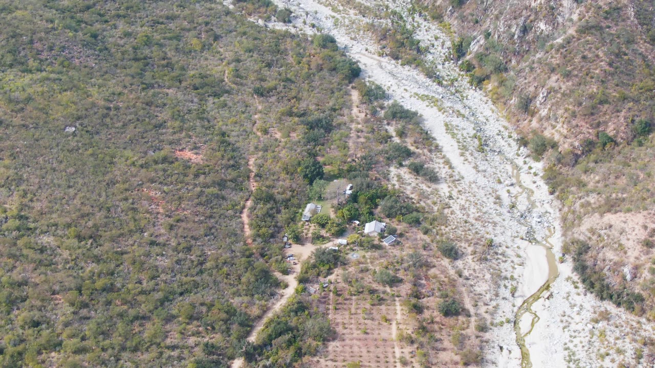 Drone view of a dry river channel curving past hillside vegetation and small buildings.