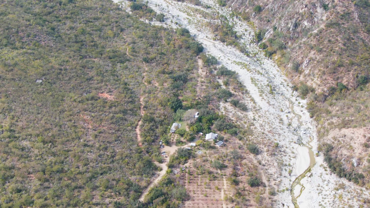 High aerial frame over San Dionisio, Baja California Sur, showing brushy mountain terrain, a pale riverbed at right, dirt access paths, a field grid at lower left, and no visible people.