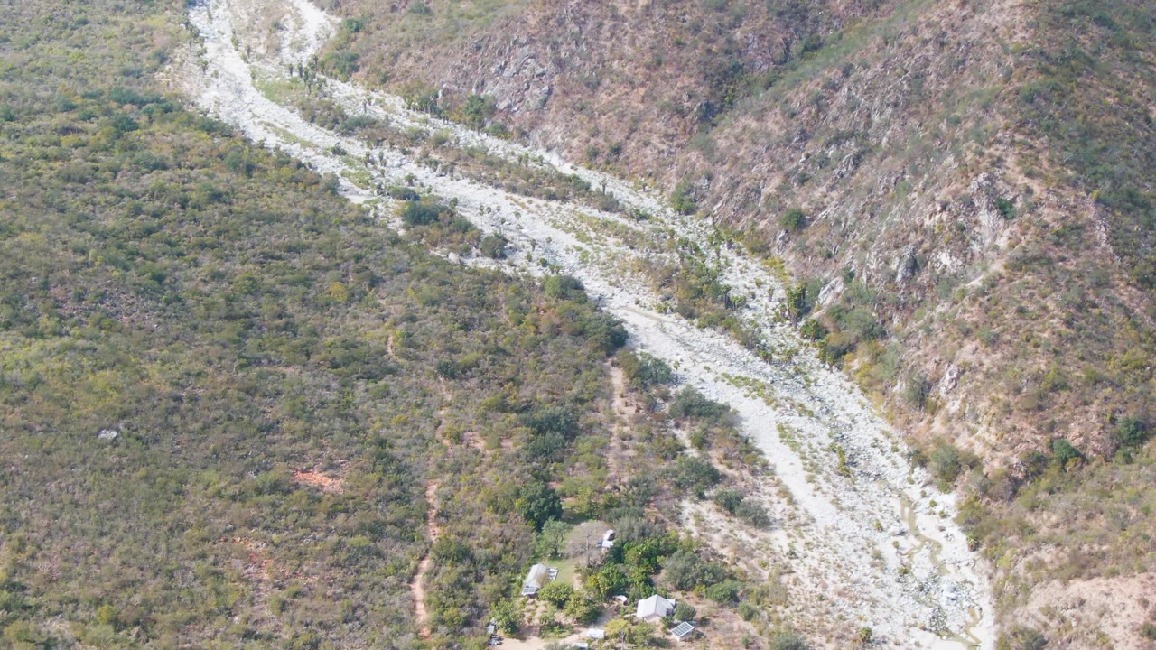 Overhead frame from San Dionisio, Baja California Sur showing a winding pale track through a dry valley with scrubby vegetation and rugged hills, with no visible people.