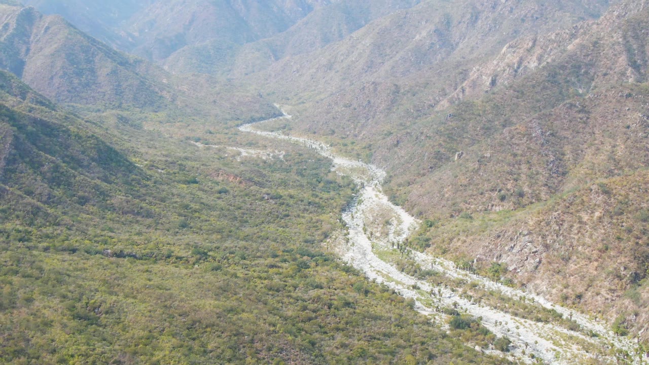 Aerial frame showing the river channel bending through dense green brush, with steep rocky slopes behind it in San Dionisio, Baja California Sur, and no visible people.