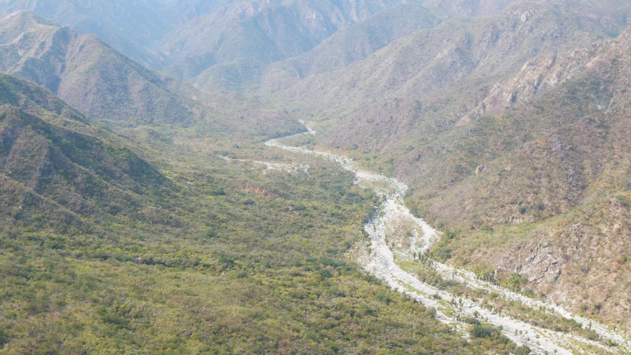Aerial view of the river curving through the lower half of the valley, with hazy mountain ridges layered behind San Dionisio, Baja California Sur, and no visible people.
