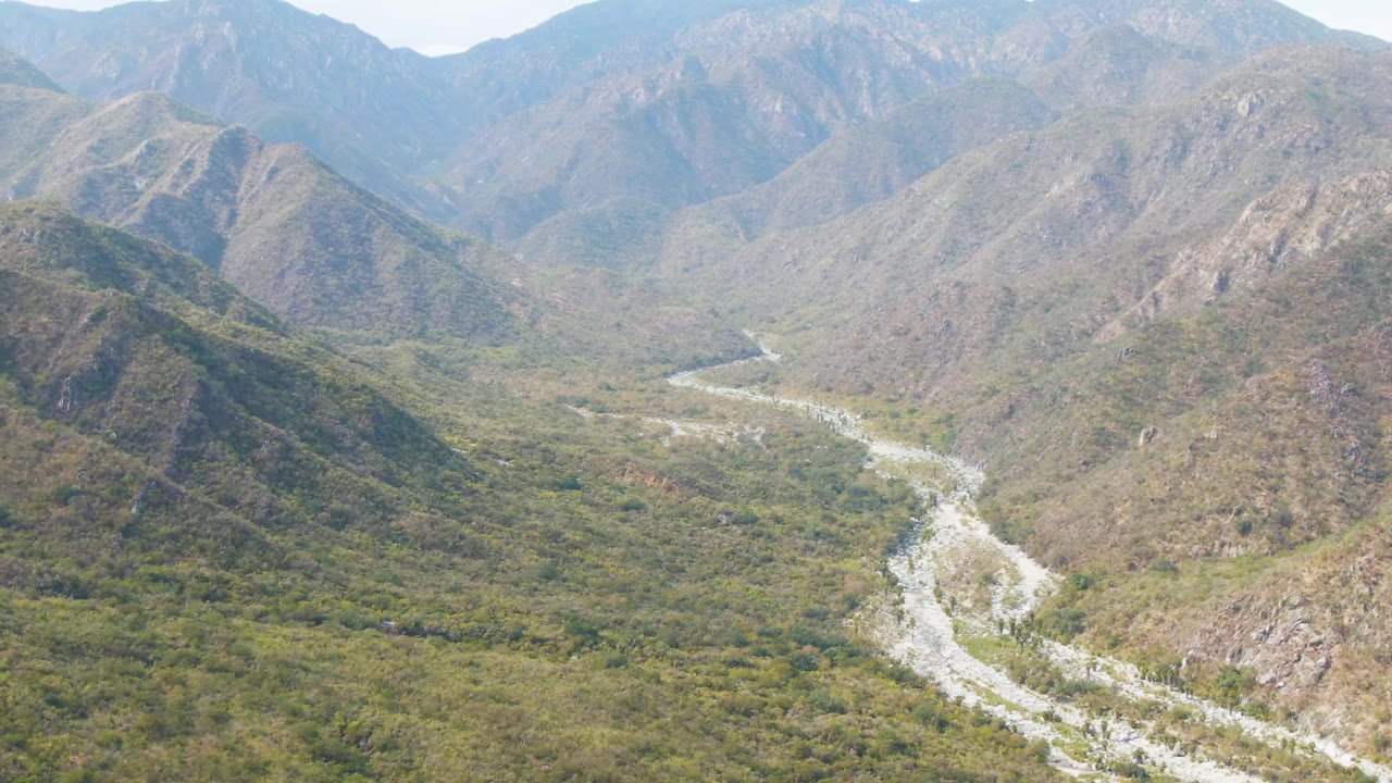 Aerial view in San Dionisio, Baja California Sur, looking over a pale winding riverbed through green and brown mountain ridges under a soft sky, with no visible people.