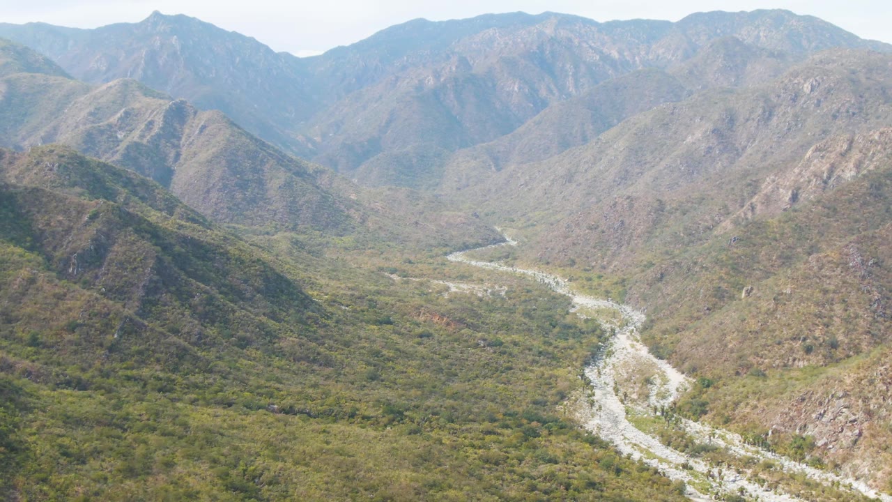 San Dionisio aerial frame with the river bending through dense valley vegetation, rugged brown-green hills on both sides, distant mountains, and no visible people.